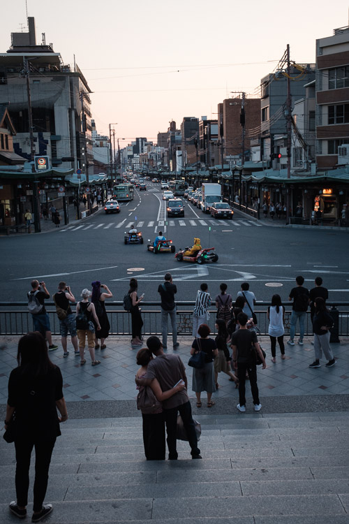 Onlookers watch from temple steps as costumed tourists drive go-karts through a wide Kyoto intersection at dusk
