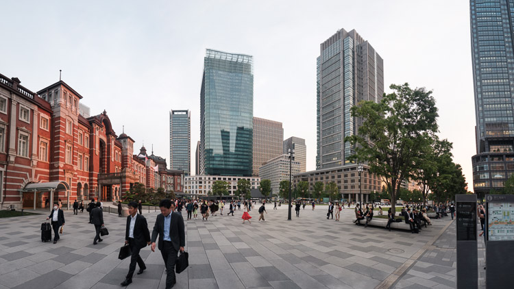 Commuters cross the broad paved plaza between the red brick facade of Tokyo Station and glass towers at Marunouchi Square