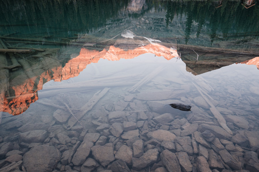 A sunlit orange peak and evergreens reflected upside down on a placid lake, submerged stones showing through the glassy water