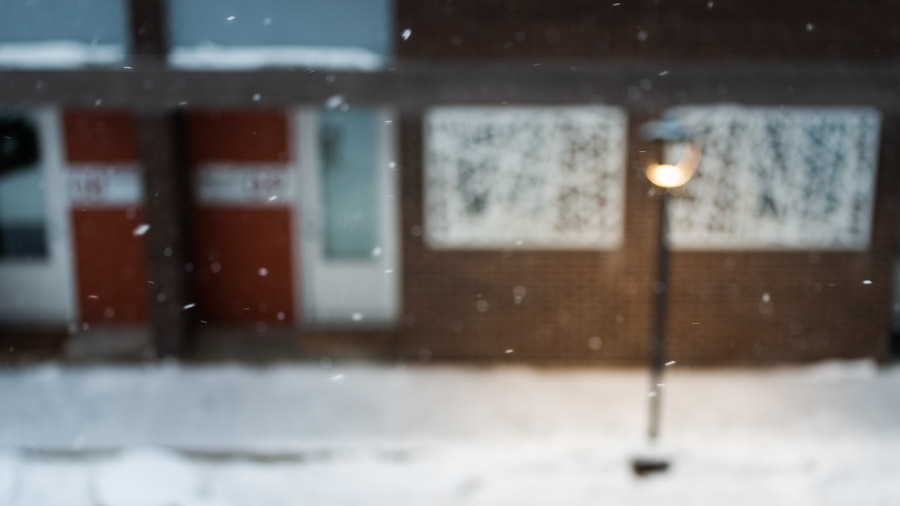 An out-of-focus streetlamp glowing warm against a brick apartment block as snow falls at dawn