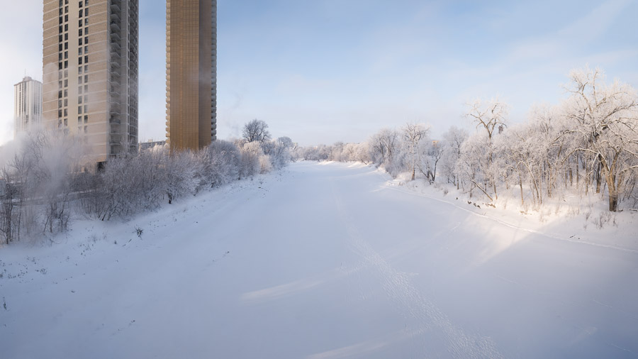 Two high-rise towers standing over the snow-covered frozen river, hoarfrosted trees lining the banks under soft blue sky