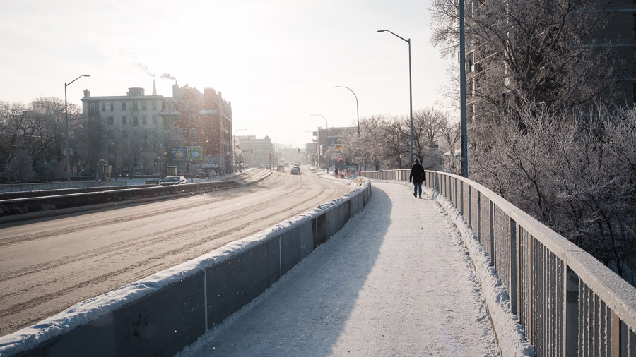 A lone figure walks the frost-dusted sidewalk of a bridge over an empty street, winter sun hazing the buildings beyond