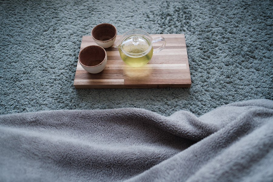 A glass teapot of green tea and two ceramic cups on a wooden board, set on a soft grey rug beside a blanket
