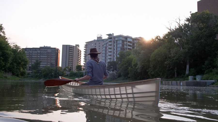 A paddler in a wide-brimmed hat steers a wooden canoe up the calm Assiniboine River toward apartment blocks at sunset