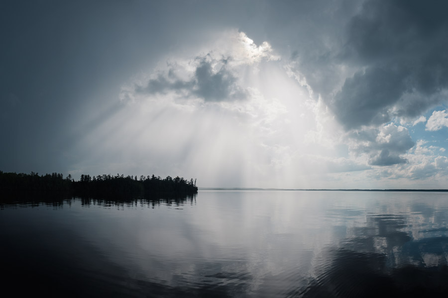 Shafts of sunlight bursting through a storm cloud over a mirror-calm lake and treed shoreline