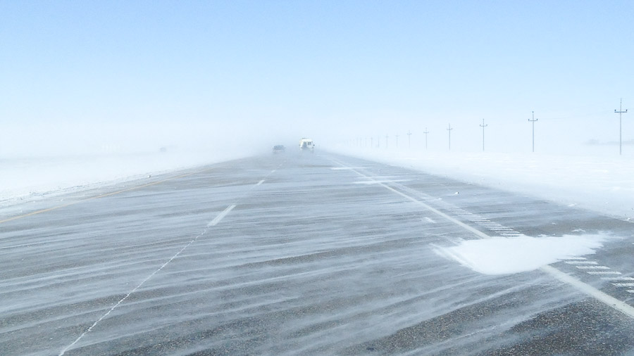 Snow drifting across a prairie highway in near-whiteout, distant vehicles and power poles fading into the blowing white