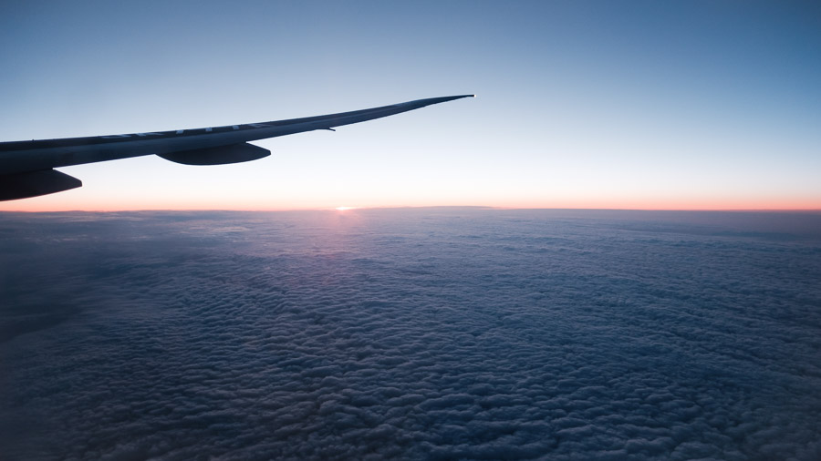 An airplane wing over a quilt of clouds at dusk, the horizon glowing orange beneath a deep blue sky