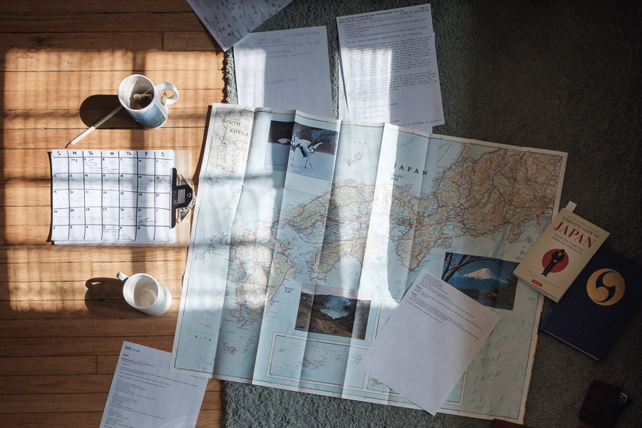 A trip-planning spread on a sunlit floor: a large map of Japan, a hand-drawn calendar, printed notes, an etiquette guidebook, and mugs of tea