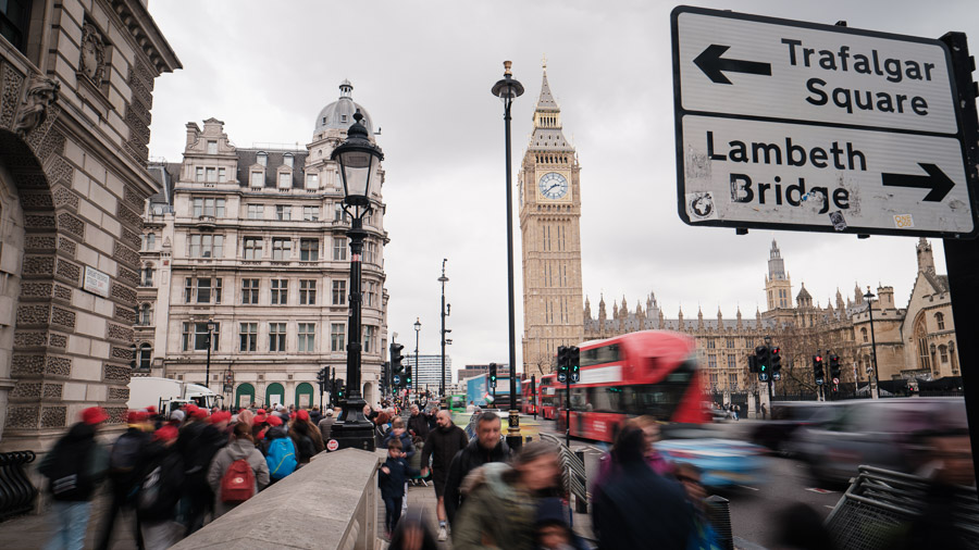 Crowds and a red double-decker bus blur past street signs for Trafalgar Square and Lambeth Bridge, Big Ben rising sharp behind