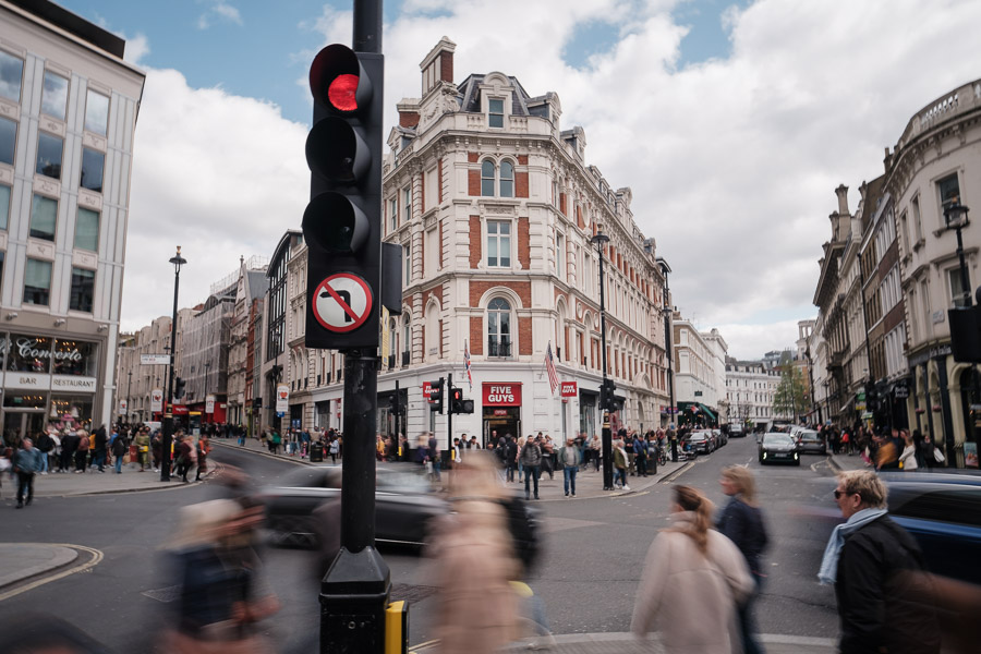 Pedestrians and a black cab blurring through a London intersection beneath a red traffic light, ornate Victorian facades behind