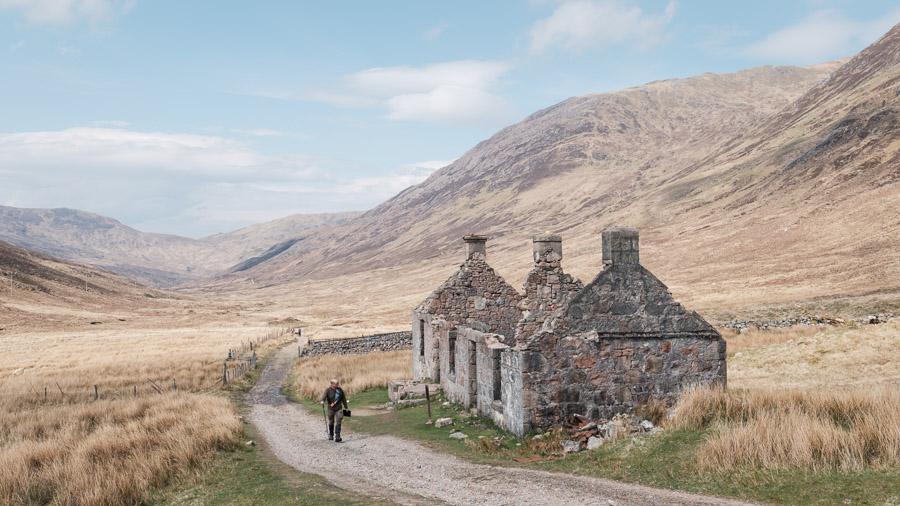 A hiker walks a gravel track past a roofless stone cottage ruin in a wide moorland valley on the West Highland Way
