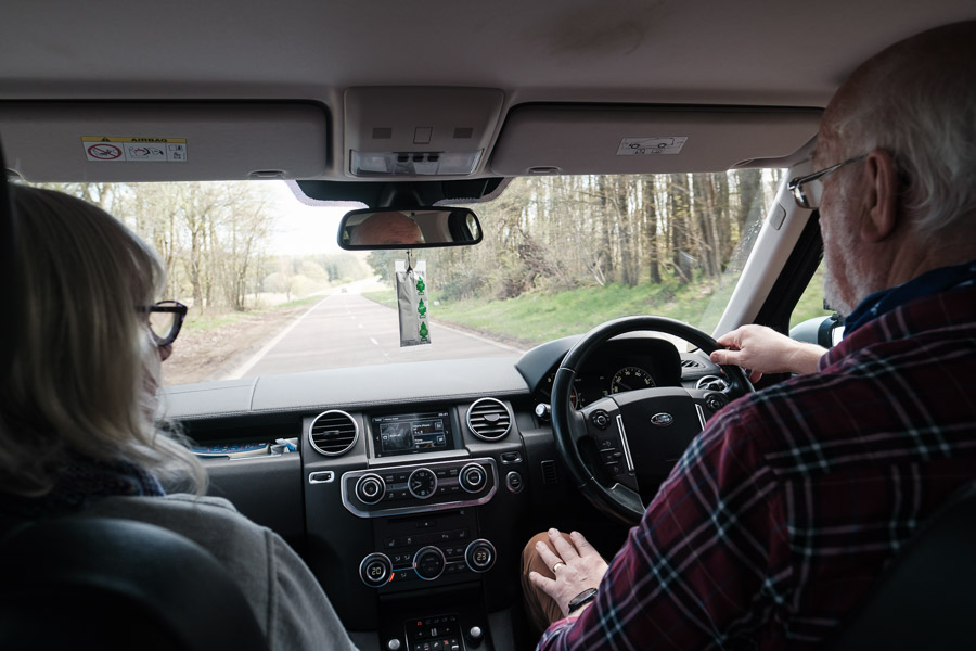 Backseat view of Allan at the right-hand wheel and Ann beside him as the car rolls down a tree-lined road