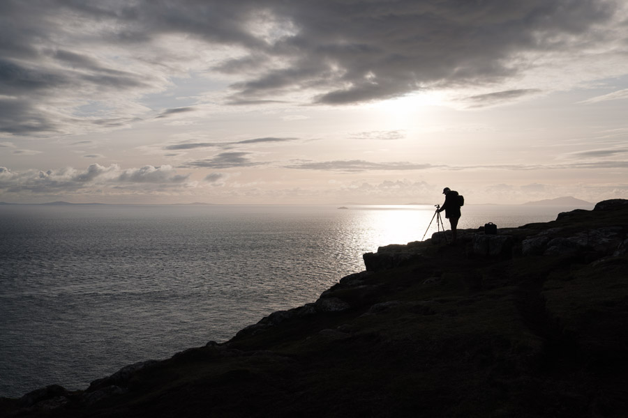 A photographer silhouetted at a tripod on a clifftop, the sea glowing silver under an evening sky