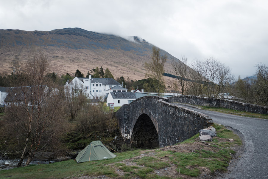 A green tent pitched by the river below the old stone arch of Bridge of Orchy, the white hotel and cloud-capped hills behind