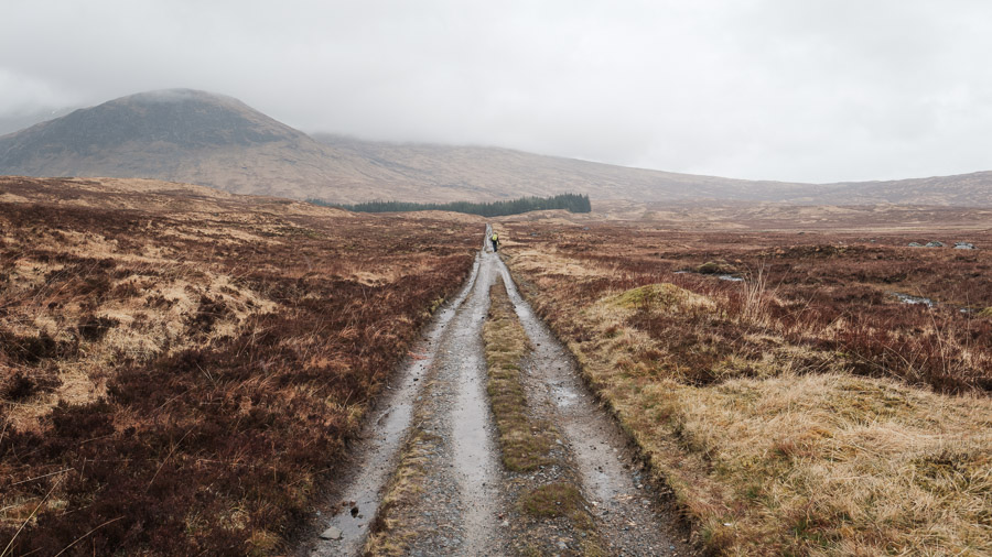 A distant hiker in rain gear follows the wet gravel of an old military road across open moorland toward mist-hidden hills