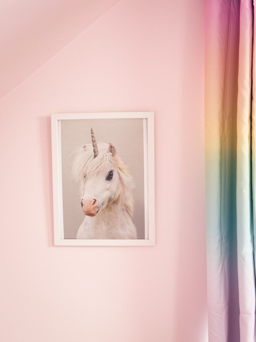 A framed portrait of a unicorn with a pastel rainbow mane on a pink bedroom wall beside rainbow curtains