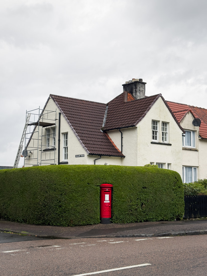 A red Royal Mail post box tucked into a neatly clipped hedge wrapping the corner of a white Scottish house