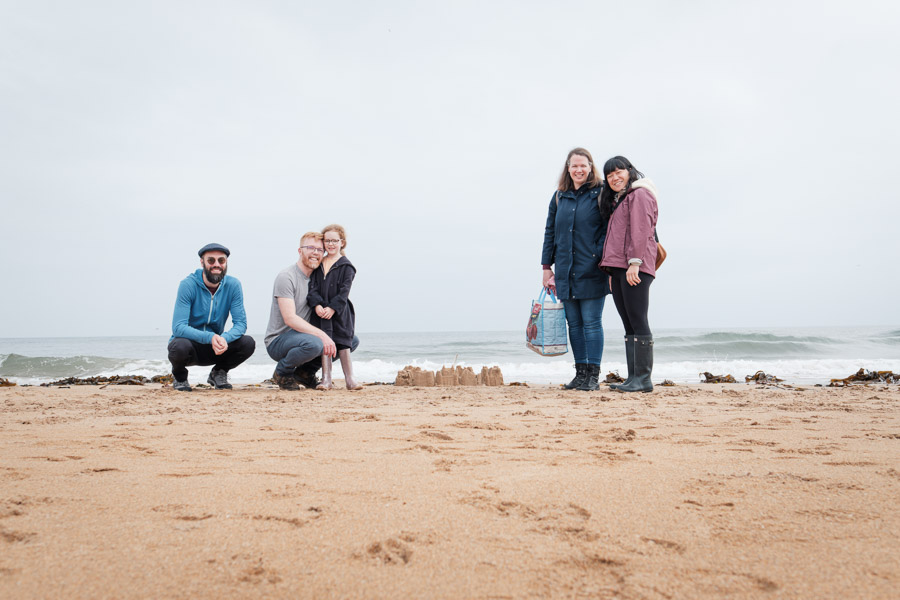 Five family members posing proudly around a sandcastle on a wide Fife beach as waves roll in behind