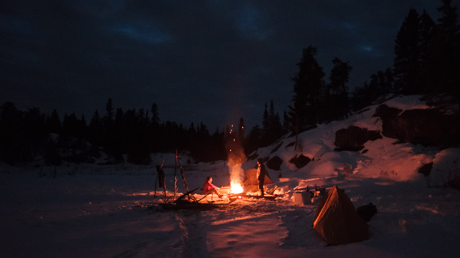 Two winter campers around a roaring bonfire on a frozen lake at dusk, sparks rising toward dark spruce and a small tent glowing orange