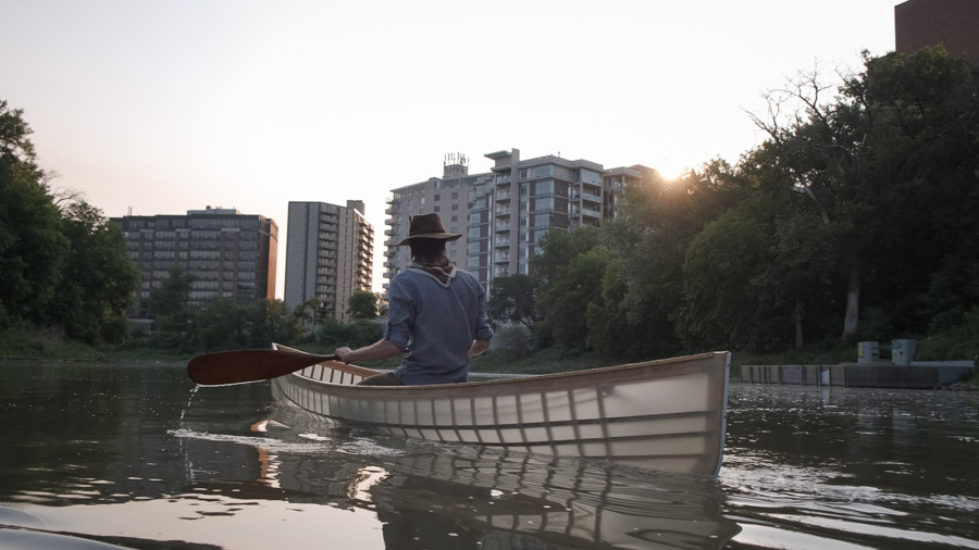 A paddler in a wide-brimmed hat steers a handmade wooden canoe up the Assiniboine River at sunrise, apartment towers rising above the trees