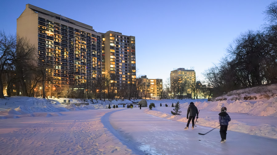 Two hockey players skate the freshly flooded river trail at dusk beneath glowing apartment towers