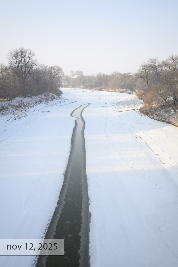 Labeled nov 12, 2025: the Assiniboine River from the Osborne Street Bridge in Winnipeg, a dark ribbon of open water winding through fresh snow