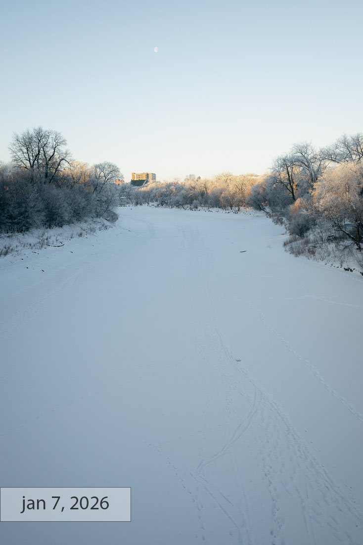 Labeled jan 7, 2026: the frozen river criss-crossed with footprints under a clear blue sky, a daytime moon overhead and low sun catching the treetops