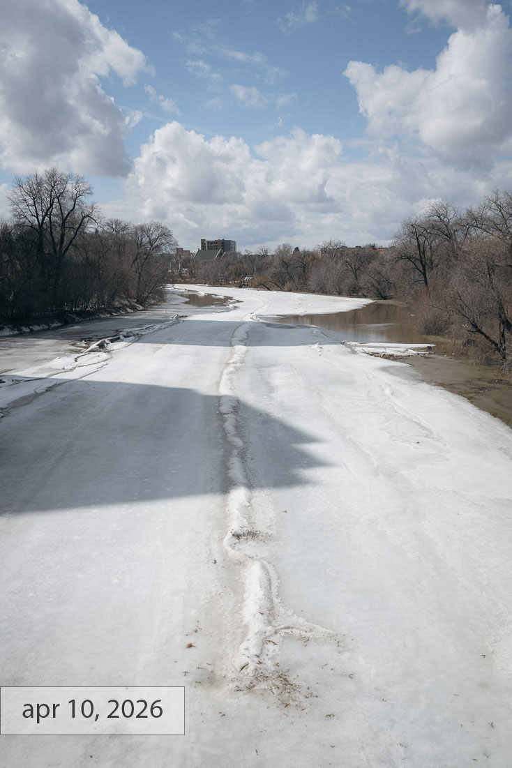 Labeled apr 10, 2026: the river ice turning grey and rotten in spring, open water appearing along the banks under bright cumulus clouds